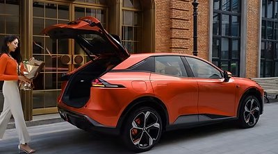 Orange electric car with open boot next to a woman carrying flowers.