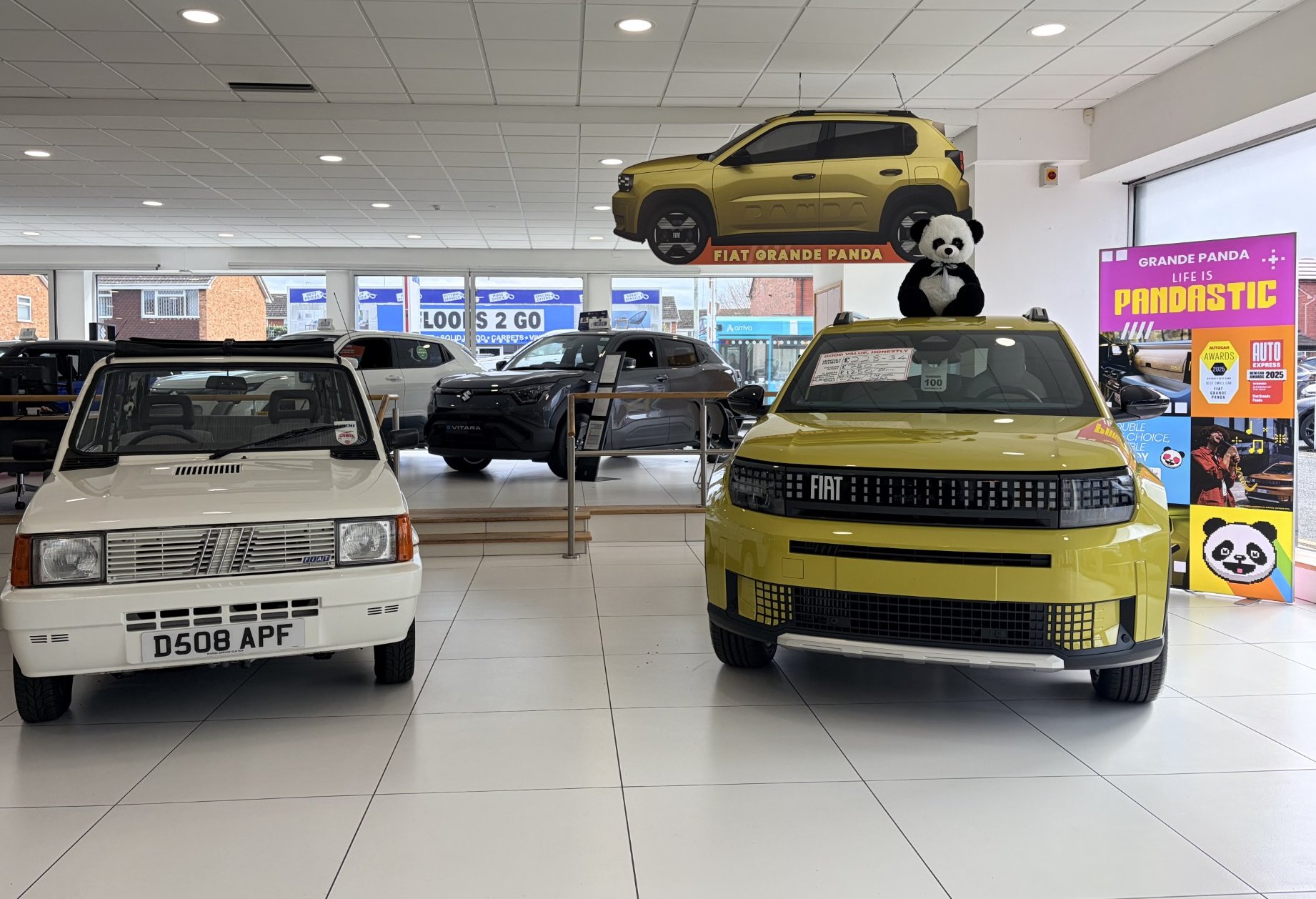 Two Fiat Panda cars, one white classic and one modern yellow, in a showroom with a panda toy.