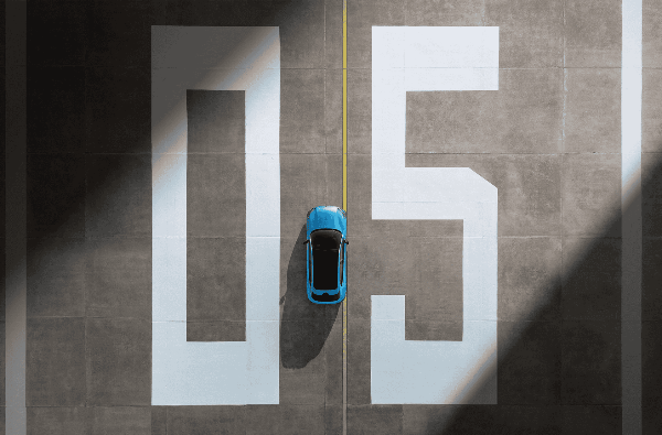 Blue car drives between large painted numbers 0 and 5 on a paved surface.